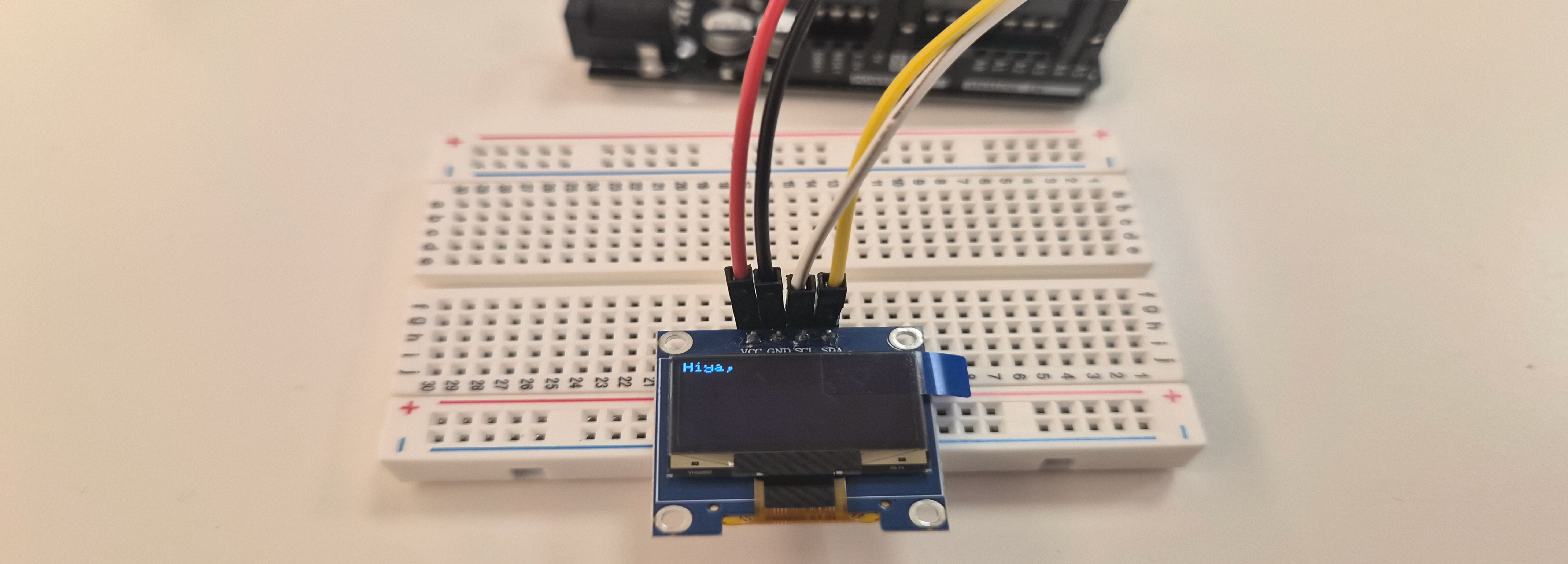 An image close-up of a breadboard with four wires attached (red, black, white and yellow) connected to a LCD that displays Hiya, 